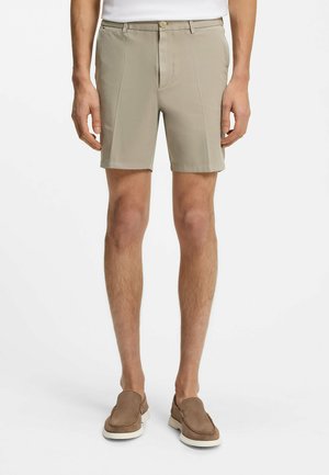 Mężczyzna ubrany w beżowe, dopasowane szorty i brązowe wsuwane buty na jednolitym białym tle, stojący z luźno opuszczonymi ramionami.