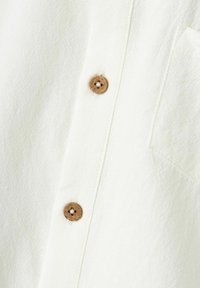 Biała koszula z teksturowanej tkaniny, z zapięciem z jasnego, drewnianego guzika oraz małą kieszonką na piersi.