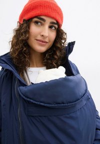 Giubbotto imbottito blu navy con una grande tasca anteriore che tiene un orsetto di peluche bianco. Il modello indossa un berretto a coste arancione brillante. Capelli ricci visibili.