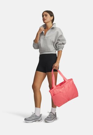 Grijze sweatvest met rits en voorzak, zwarte korte broek, grijze sneakers, en een felroze draagtas met dubbele handgrepen.