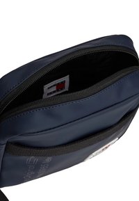 Granatowa torebka crossbody o gładkiej, błyszczącej teksturze. Zawiera zamek błyskawiczny i czarny pasek, a wewnątrz znajduje się widoczne logo i metka.