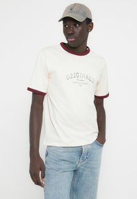 Krém színű póló bordó szegéllyel, közepén egy merész "ORIGINALS" logóval. Világoskék farmernadrággal párosítva, laza megjelenésért.