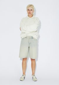 Jasnozielona, oversize koszulka polo, zestawiona z luźnymi, jasnoniebieskimi szortami dżinsowymi oraz białymi sneakersami. Prosty krój; delikatne postrzępienia na krawędzi szortów.