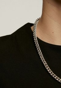 Collar de cadena de plata con eslabones gruesos entrelazados, posado sobre una camiseta negra de manga corta; textura suave y acabado pulido.