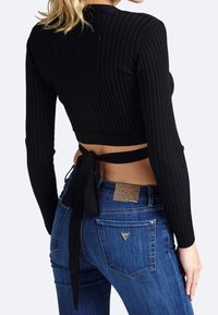 Donna che indossa un crop top a maniche lunghe nero a costine con un nodo sulla schiena, abbinato a jeans in denim blu, vista da dietro.