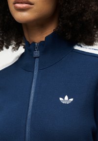 Gros plan d'une personne portant une veste zippée bleu marine avec un col côtelé et le logo blanc Adidas trefoil sur la poitrine.