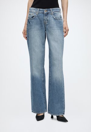 Persona che indossa jeans a vita alta, azzurri a gamba larga e scarpe nere a punta, in piedi contro uno sfondo semplice. Solo la parte inferiore del busto è visibile.
