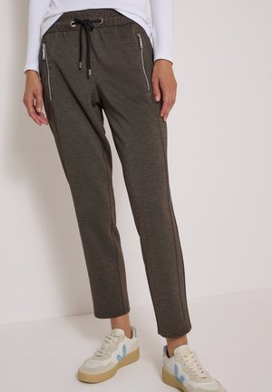 Femme portant un pantalon gris foncé à cordon avec des poches latérales zippées et des baskets blanches avec des détails bleu clair, debout contre un mur uni.