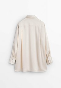 Blouse beige à manches longues avec un pli au dos, des poignets boutonnés et un col classique suspendue sur un fond blanc.