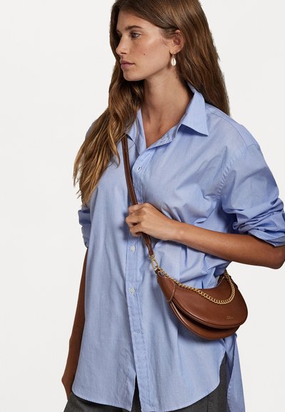 Ruda odinė petnešėlė su iškiliu pavidalu ir grandinėlės akcentu, dėvima per kūną ant šviesiai mėlynos marškinėlių su mygtukais. Lygus paviršius.