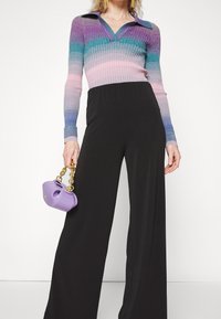 Personne portant un pull côtelé multicolore avec col et un pantalon noir à jambes larges, tenant un petit sac à main violet avec une anse chaîne dorée.