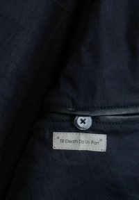 Material textil negru cu o textură netedă, având o etichetă cusută pe care scrie „Până la moarte să ne despartă”, fixată cu un nasture rotund argintiu.