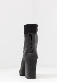 Bota negra de tacón alto con acabado brillante de charol, cuello tejido texturizado y cremallera lateral para el cierre. La suela de goma proporciona agarre.