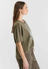 Olijfgroene t-shirt met korte mouwen en een relaxte pasvorm, voorzien van een grafische print op de voorkant, gecombineerd met corduroy broek in een vergelijkbare kleur.