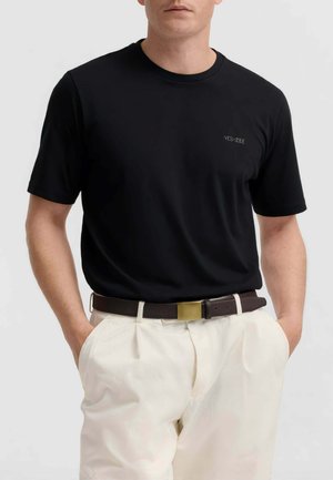 Uomo che indossa T-shirt nera a maniche corte con piccolo logo sul petto, pantaloni bianchi plissettati e cintura marrone scuro con fibbia dorata.