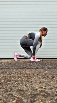 Woman in gray leggings and white-pink sneakers tying shoelaces, crouched on textured ground in front of white garage door.
