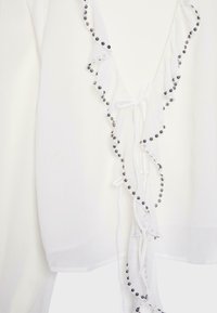 Vêtement blanc avec des bords froncés orné de petites perles métalliques et d'une fermeture à nouer à l'avant.