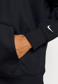 Sudadera negra de Nike con puños acanalados, bolsillo lateral y pequeño logotipo blanco en la manga. Textura de tela suave, diseño casual.