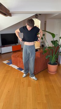 Pantalon plissé gris à coupe décontractée, associé à une chemise noire à manches courtes. Chaussures de sport argentées. Intérieur avec des plantes.