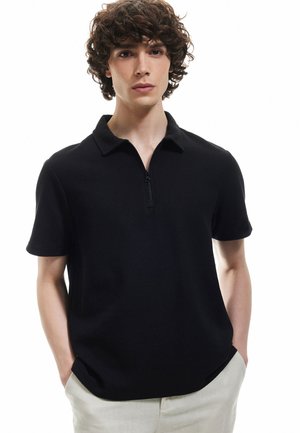 Camisa polo negra con cremallera, mangas cortas y tejido texturizado. El modelo posa con las manos en los bolsillos, sobre un fondo neutro.