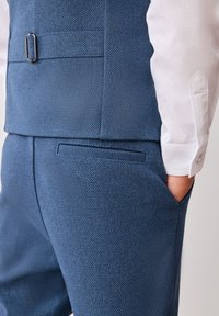 Traje azul texturizado con un chaleco ajustado y un patrón sutil. Presenta un regulador lateral y un bolsillo trasero. Acompañado de una camisa blanca.