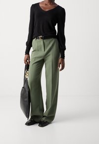 Maglione nero a scollo a V, pantaloni verdi a vita alta con vestibilità rilassata, cintura nera con accessori dorati e borsa tote nera.