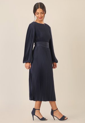 Navy pleated midi dress with long, puff sleeves and a belted waist; paired with navy high-heeled sandals featuring ankle straps.