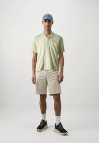 Kurzärmeliges, hellgrünes Hemd mit Knöpfen und Tasche, kombiniert mit beigen Shorts. Getragen mit einer hellblauen Mütze und schwarzen Turnschuhen.