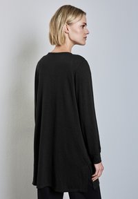 Musta pitkähihaisen paita, jossa on rento mitoitus, sivuaukot ja pehmeä rakenne; pyöreä kaula-aukko ja hieman laskeutuvat hihat, yksinkertainen muotoilu.
