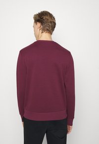 Sudadera color marrón oscuro con mangas largas, escote redondeado y puños acanalados. Textura suave con una cintura ajustada y sin logos ni patrones visibles.