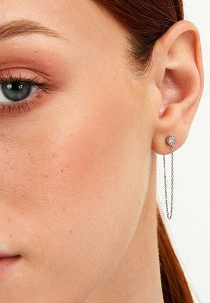 Sølv stud ørestik med en rund, klar sten og en sart kæde, der strækker sig nedad. Huden omkring øret er synlig.