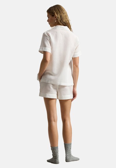 Vrouw staand met de rug naar voren, gekleed in een wit T-shirt met korte mouwen, bijpassende korte broek en grijze gebreide sokken op een effen achtergrond.