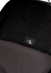 Čierny látkový batoh s hladkým interiérom, obsahuje sieťovanú kapsu a štítok s nápisom "CK". Zapínanie na zips.