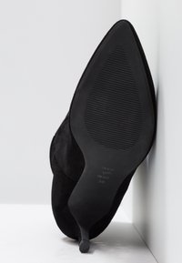 Chaussure à talon haut en daim noir à bout pointu avec semelle en caoutchouc texturée et talon fin, taille 38, placée debout contre un mur d'angle.