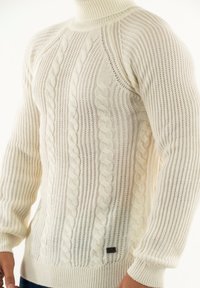 Jersey blanco de punto con una textura de canalé, patrón de trenzado, cuello alto y mangas ajustadas, con una pequeña etiqueta de la marca en el dobladillo.