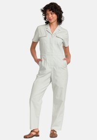 Lichtgrijze korte mouwen jumpsuit met een kraag, twee borstzakken en een ontspannen pasvorm. Zacht materiaal met zichtbare naden.