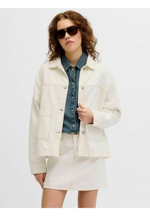 Noor naine lokkis juustega kannab tumedaid päikeseprille, valget jopet, teksasärki ja valget seelikut, seisab tavalise tausta ees.