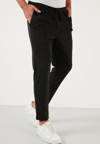 Černé sportovní tepláky s elastickým pasem, bočními kapsami, strukturovanou tkaninou a zúženými nohavicemi, doplněné bílými teniskami.