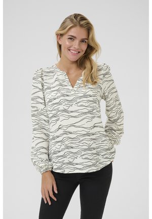 Blouse blanche avec des rayures noires ondulées, décolleté en V, et manches longues avec des poignets froncés. Associée à un pantalon noir ajusté.