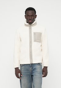 Veste en polaire crème avec des accents gris, col montant, fermeture éclair sur toute la longueur à l'avant et poches à l'avant. Arrière-plan texturé subtil et marquage sur la poche.