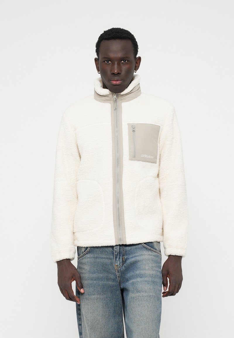 Veste en polaire crème avec des accents gris, col montant, fermeture éclair sur toute la longueur à l'avant et poches à l'avant. Arrière-plan texturé subtil et marquage sur la poche.