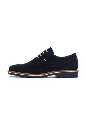 Zapato de ante azul marino con una textura suave, cordones crema, un logo sutil en el lateral y una suela de color marrón claro en contraste.
