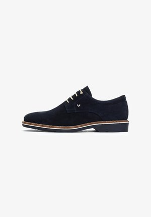 Zapato de ante azul marino con una textura suave, cordones crema, un logo sutil en el lateral y una suela de color marrón claro en contraste.