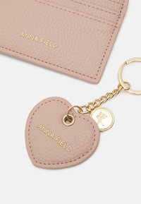 Pink leather keychain in heart shape with gold hardware. Features embossed brand name "ANNA FIELD" and decorative charm attached by a chain.