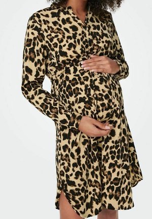 Gravid kvinde iført en langærmet kjole med leopardprint, der holder om sin mave med begge hænder mod en ensartet hvid baggrund.