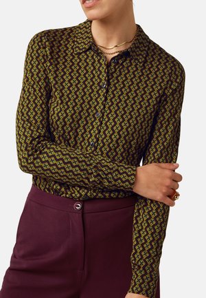 Vrouw draagt een lange mouwen patroonoverhemd met geometrische vormen en hooggetailleerde donkerpaarse broek, draagt gouden sieraden.