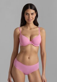 Νεαρή γυναίκα με μακριά σκούρα μαλλιά φοράει σετ ροζ σουτιέν και σλιπ, στεκόμενη μπροστά σε απλό γκρι φόντο.