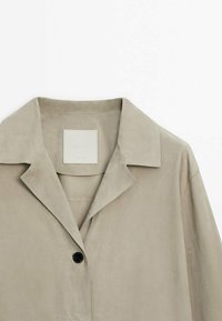 Vaaleanbeige, mokkanahkainen takki, jossa on yksi musta nappi ja neliömäinen etiketti sisäpuolella. Silkkinen pinta ja yksinkertainen muotoilu.