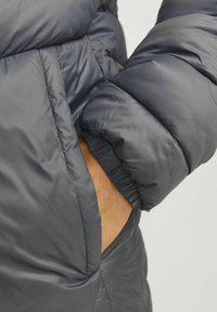 Chaqueta de plumas en gris oscuro, con un diseño acolchado, puños elásticos y un bolsillo lateral. La tela tiene una textura suave y brillante.