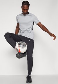 Γκρι αθλητικό μπλουζάκι με μαύρες και κίτρινες λεπτομέρειες, μαύρο παντελόνι και μαύρα ποδοσφαιρικά παπούτσια. Μπάλα με γεωμετρικά σχέδια και κόκκινο λογότυπο.